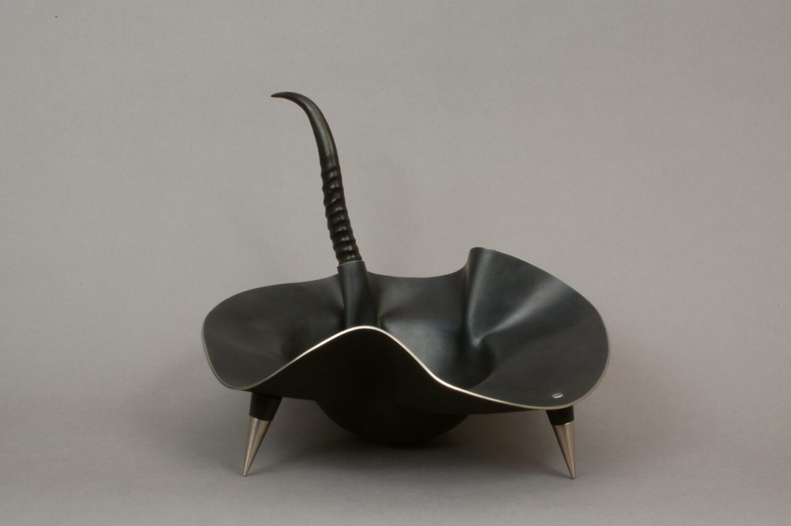 A black bowl with metal legs on a gray background, perfect for displaying Ira Sherman Jewelry.